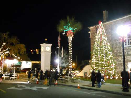 A festive street scene at night, featuring a decorated tree, clock tower, and palm trees adorned with holiday lights.