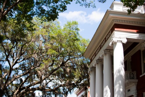 Historic building with white columns, surrounded by lush green trees and Spanish moss under a blue sky.
