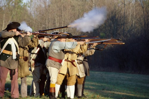A group of historical reenactors firing muskets in a wooded area, smoke rising from the barrels.