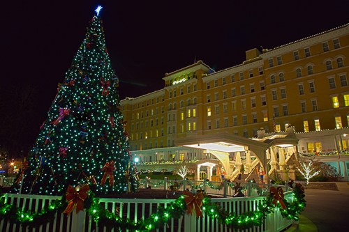A brightly lit Christmas tree adorned with decorations stands in front of a large, illuminated hotel at night.