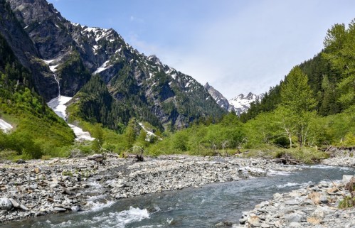 A serene mountain landscape with a river, rocky terrain, and snow-capped peaks under a clear blue sky.