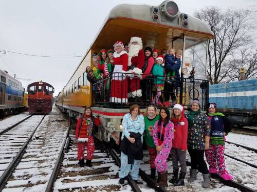 A festive group in holiday attire poses on a train car, with Santa and children, surrounded by snow and other trains.