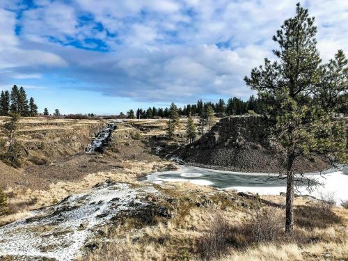 A scenic landscape featuring a frozen pond, rocky terrain, and pine trees under a partly cloudy sky.