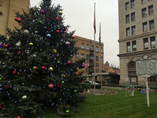 A decorated Christmas tree with colorful ornaments stands in a park near historic buildings and flags.
