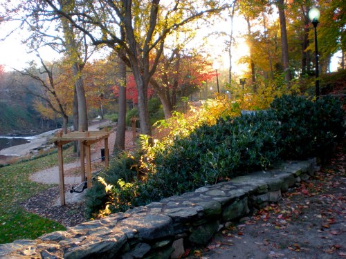 A serene park scene with colorful autumn leaves, a stone wall, and sunlight filtering through the trees.