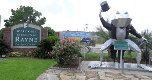 Welcome sign for Rayne, Louisiana, featuring a large frog statue in a top hat and colorful flowers.