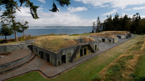 Historic concrete structures with grassy roofs, overlooking a calm body of water and surrounded by trees under a cloudy sky.
