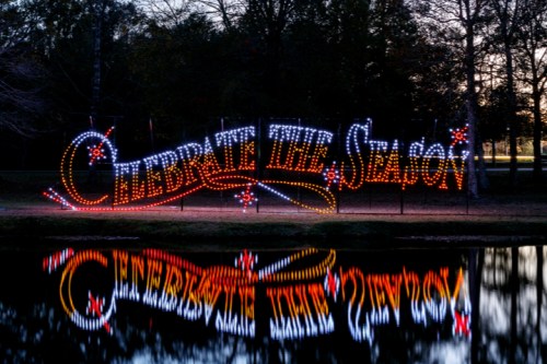 Colorful lights spell out "Celebrate the Season," reflecting in a calm pond surrounded by trees.