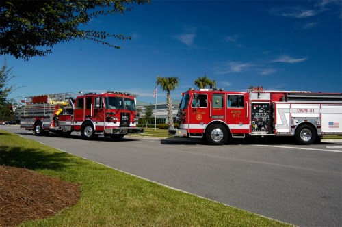 Two red fire trucks parked on a road, with palm trees and a blue sky in the background.