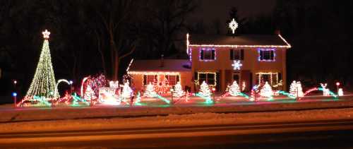 A house decorated with colorful Christmas lights, featuring a lit tree and festive displays in a snowy setting.