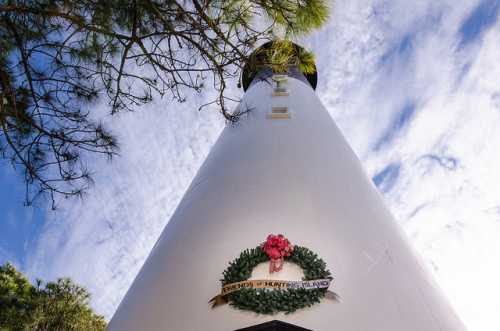 A tall lighthouse with a festive wreath at its base, surrounded by trees and a blue sky with clouds.