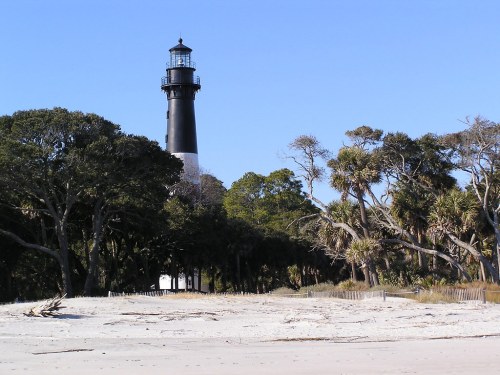 A black and white lighthouse stands among trees near a sandy beach under a clear blue sky.