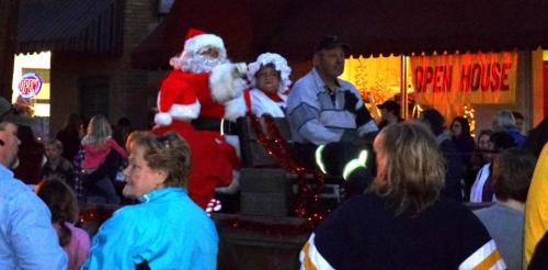 A crowd gathers as Santa and Mrs. Claus sit on a float during a festive event, with holiday decorations in the background.