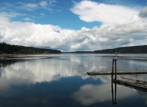 A serene lake reflects fluffy clouds and trees, with a wooden dock extending into the calm water.