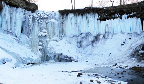 Frozen waterfall surrounded by icy cliffs and snow-covered ground, with trees in the background.