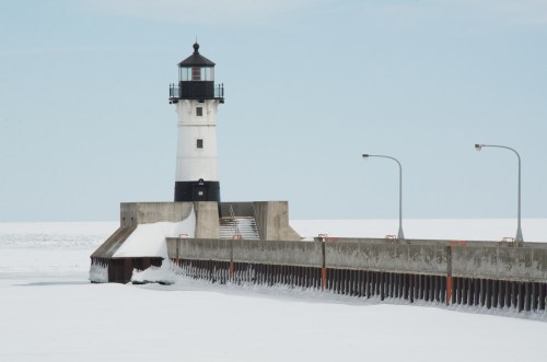 A lighthouse stands on a snowy pier, surrounded by a frozen lake under a clear blue sky.
