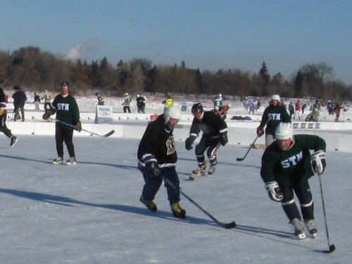 A group of people playing ice hockey on an outdoor rink, with several skaters and a clear blue sky in the background.