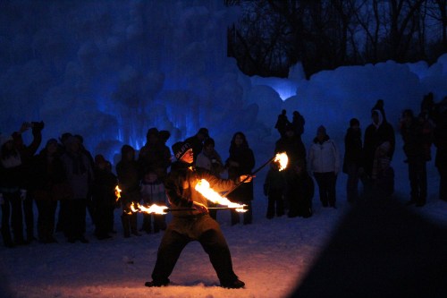 A performer twirls flaming poi in front of an audience at a winter festival, surrounded by ice formations and blue lights.