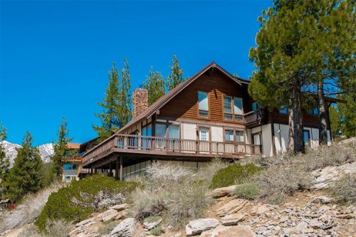A large wooden house with a deck, surrounded by trees and rocky terrain under a clear blue sky.
