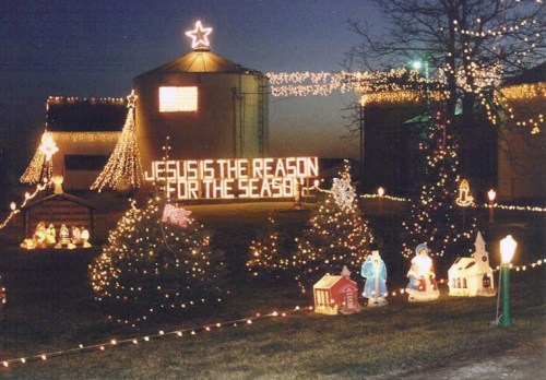 A festive outdoor scene with Christmas lights, decorated trees, and a sign reading "Jesus is the reason for the season."