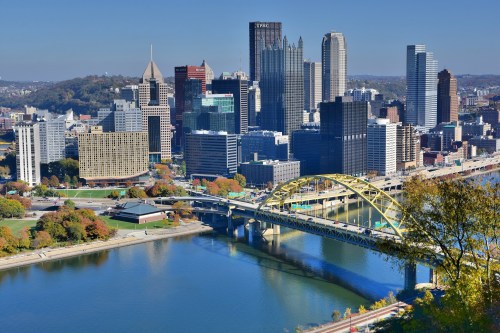 Aerial view of Pittsburgh's skyline with skyscrapers and the yellow Fort Duquesne Bridge over the river. Autumn foliage visible.