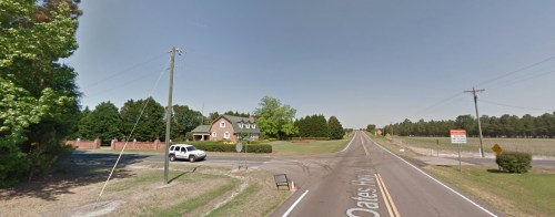 A rural road intersection with a house, trees, and a white vehicle parked nearby. Clear blue sky above.