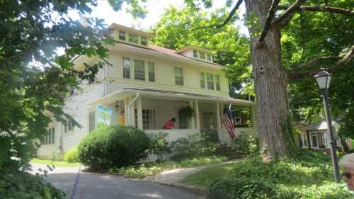 A two-story yellow house with a porch, surrounded by greenery and a large tree, featuring an American flag.