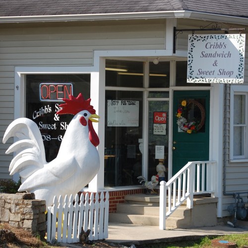 A large white rooster statue stands outside Cribb's Sandwich & Sweet Shop, which has an open sign and a welcoming entrance.
