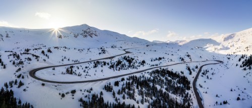 A winding road through a snowy landscape, surrounded by mountains and evergreen trees under a clear blue sky.
