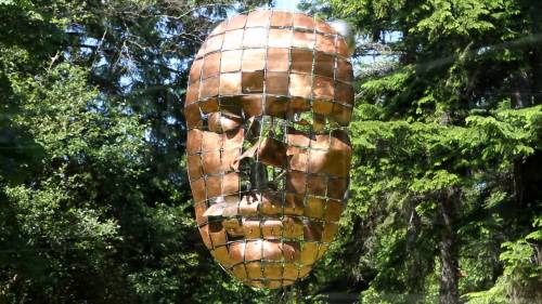 A large, reflective sculpture of a face made of metal grids, suspended among green trees in a natural setting.