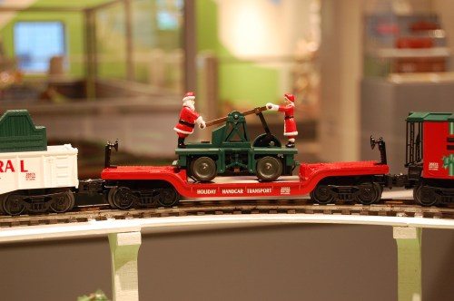 A model train with Santa figures operating a green machine on a red flatcar, surrounded by festive decorations.