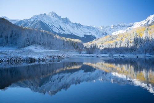Snow-covered mountains reflect in a calm lake, surrounded by autumn-colored trees under a clear blue sky.