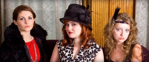 Three women in vintage attire pose together, showcasing elegant hats and stylish outfits against a decorative backdrop.