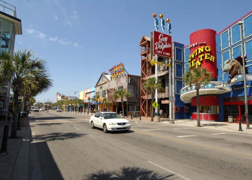 A sunny street scene featuring colorful shops and palm trees, with a car driving by in a coastal area.