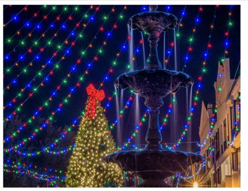A festive scene featuring a fountain and a large Christmas tree adorned with colorful lights and a red bow.