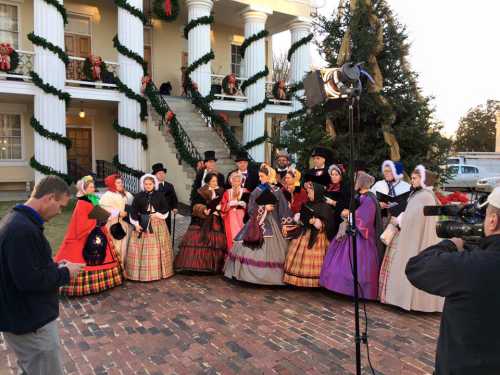 A group in historical costumes sings carols outside a decorated building during the holiday season.