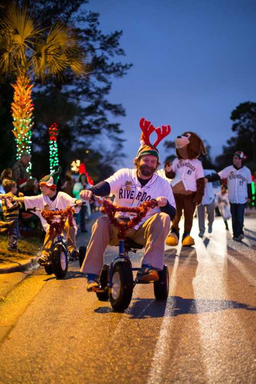 A festive parade scene with participants on tricycles, wearing reindeer antlers and holiday decorations, under evening lights.