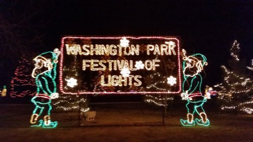 Brightly lit sign reading "Washington Park Festival of Lights," flanked by two festive elves in a nighttime setting.