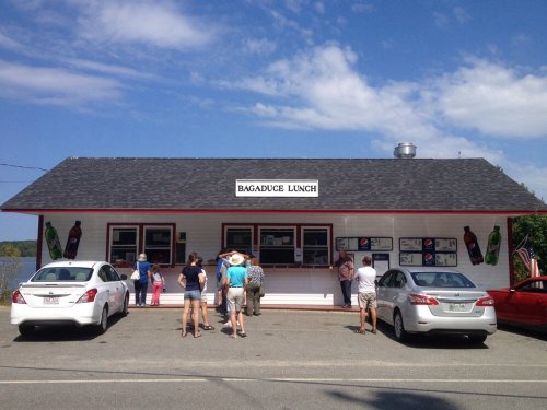 A small, white building with a black roof, featuring a sign that reads "Bagaduce Lunch," with people ordering outside.