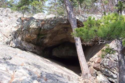 A rocky cave entrance surrounded by trees and boulders in a natural outdoor setting.