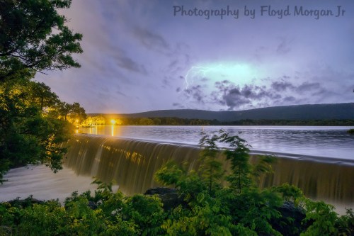 A serene waterfall at dusk, illuminated by distant lights and a faint lightning glow in the cloudy sky.