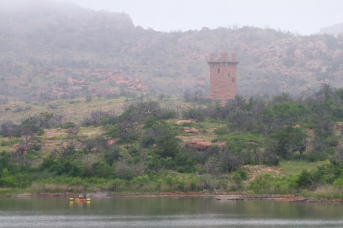 A stone tower on a hillside near a calm lake, with a kayaker paddling in the foreground and misty mountains in the background.