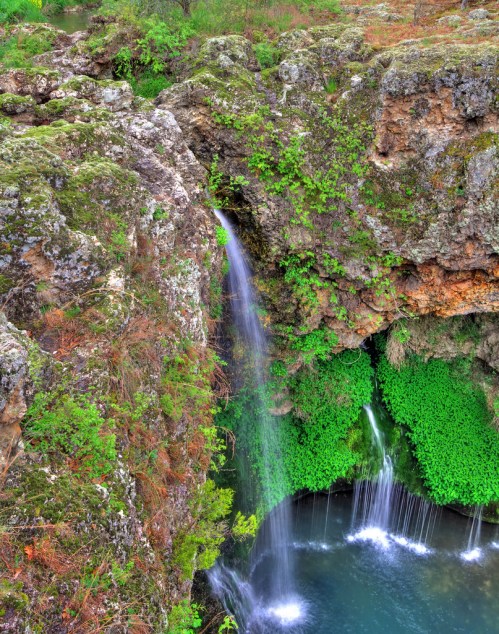 A serene waterfall cascading into a lush green pool, surrounded by rocky cliffs and vibrant vegetation.