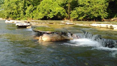 A serene river scene with smooth rocks and gentle waterfalls, surrounded by lush green trees.