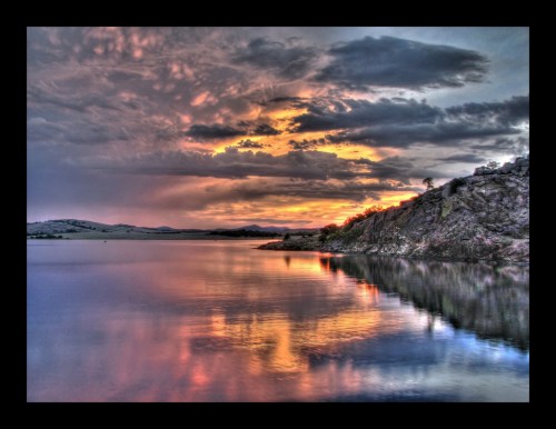 A serene lake at sunset, reflecting vibrant colors and dramatic clouds in the sky, surrounded by hills.