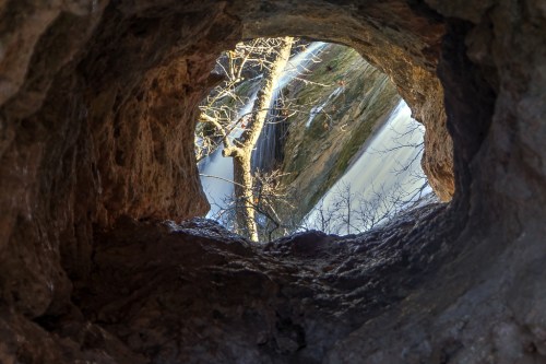 View from inside a cave looking out at a river and trees, with sunlight illuminating the scene.