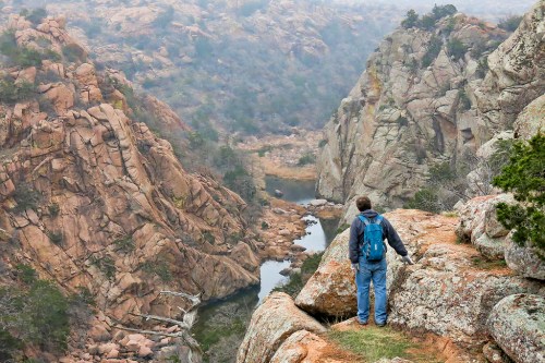 A person stands on a rocky ledge overlooking a canyon with a winding river below, surrounded by rugged terrain.