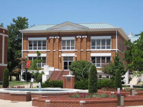 Historic brick building with a green roof, surrounded by landscaped gardens and a fountain in the foreground.