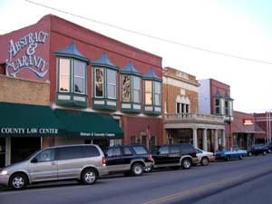 A street view of a small town featuring brick buildings, shops, and parked cars along the road.