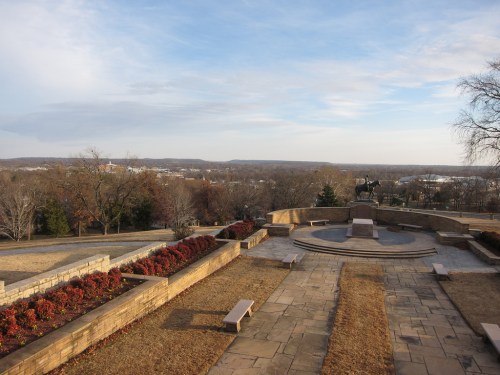 A scenic view from a park with stone pathways, flower beds, and a statue overlooking a valley and distant hills.